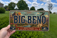 A person is holding a license plate that says "BIG BEND" with a scenic illustration of the area on it.
